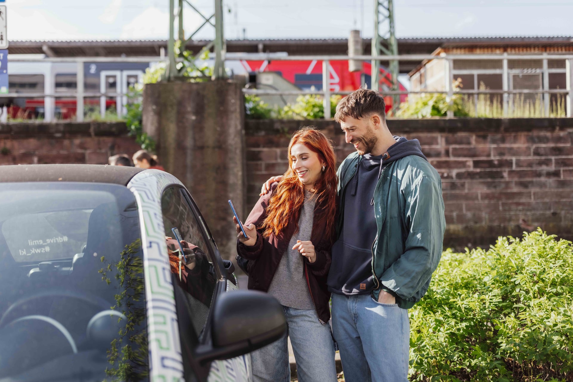 Pärchen steht am Auto, im Hintergrund ist ein Bahnhof